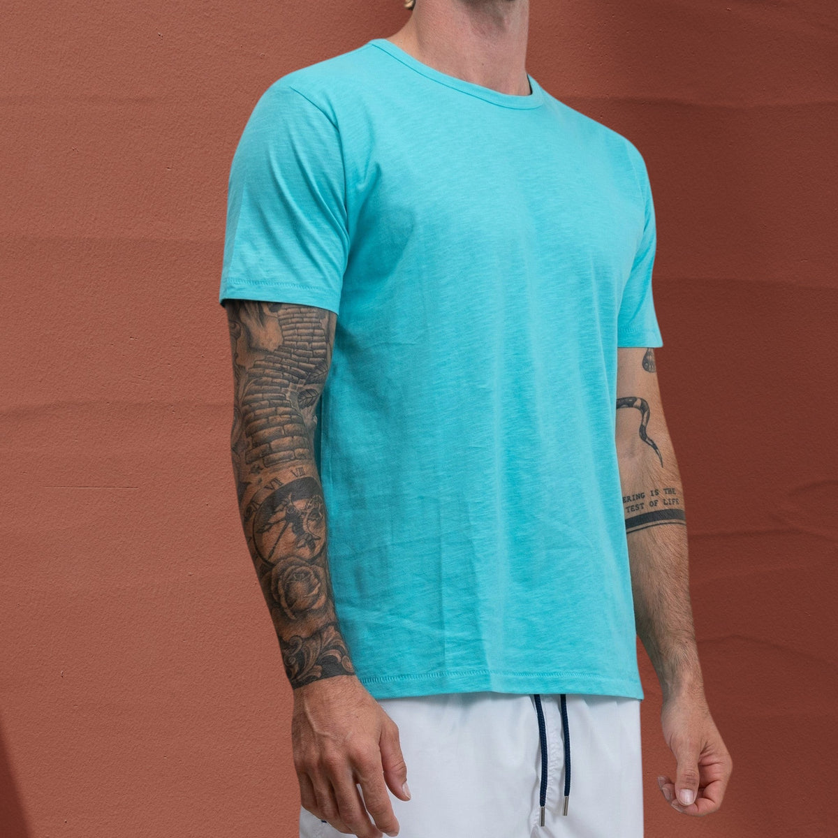 Homme portant un t-shirt St Barth French West Indies Jasper bleu turquoise en coton flammé devant un mur orange