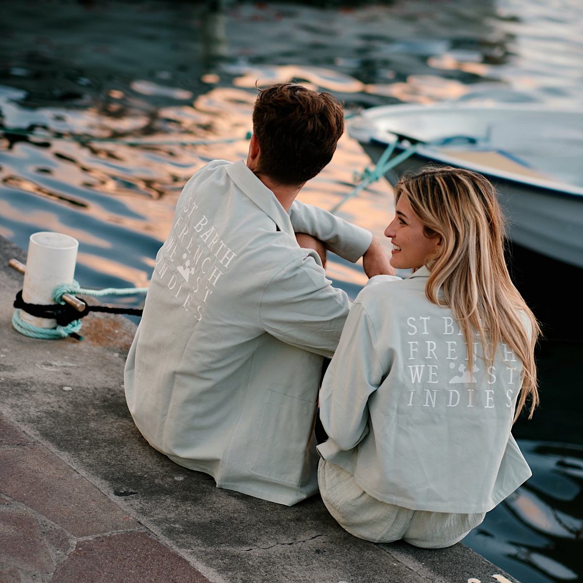 Couple portant des vestes St Barth French West Indies en sergé de coton beige avec broderie au dos près d'un port