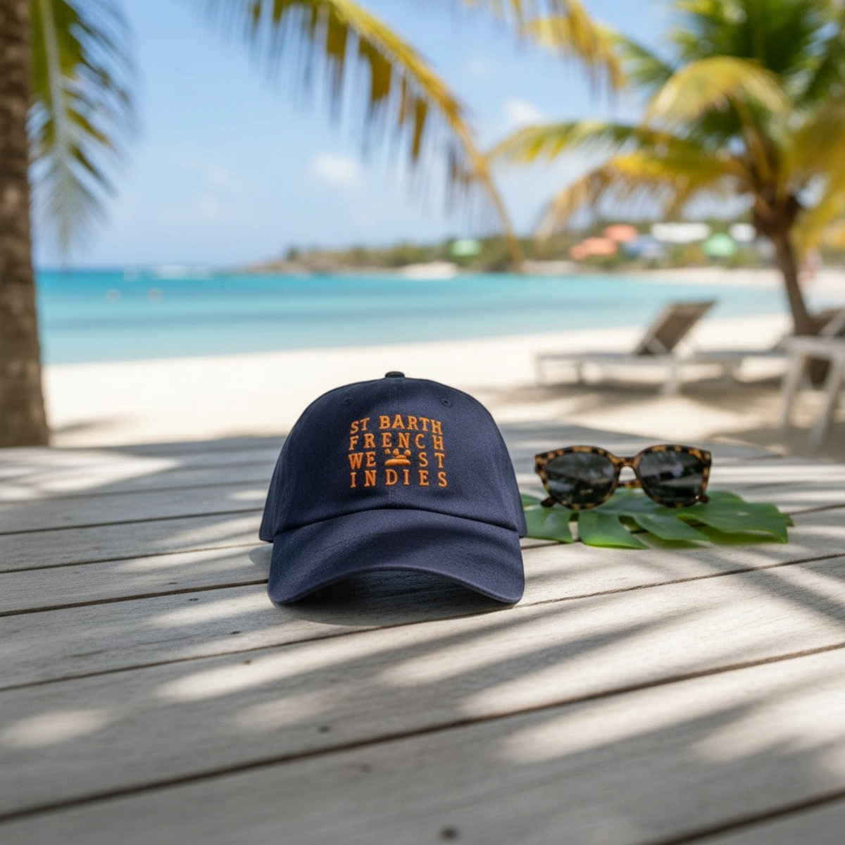 Casquette bleu marine St Barth French West Indies avec broderie orange sur une table en bois face à une plage tropicale