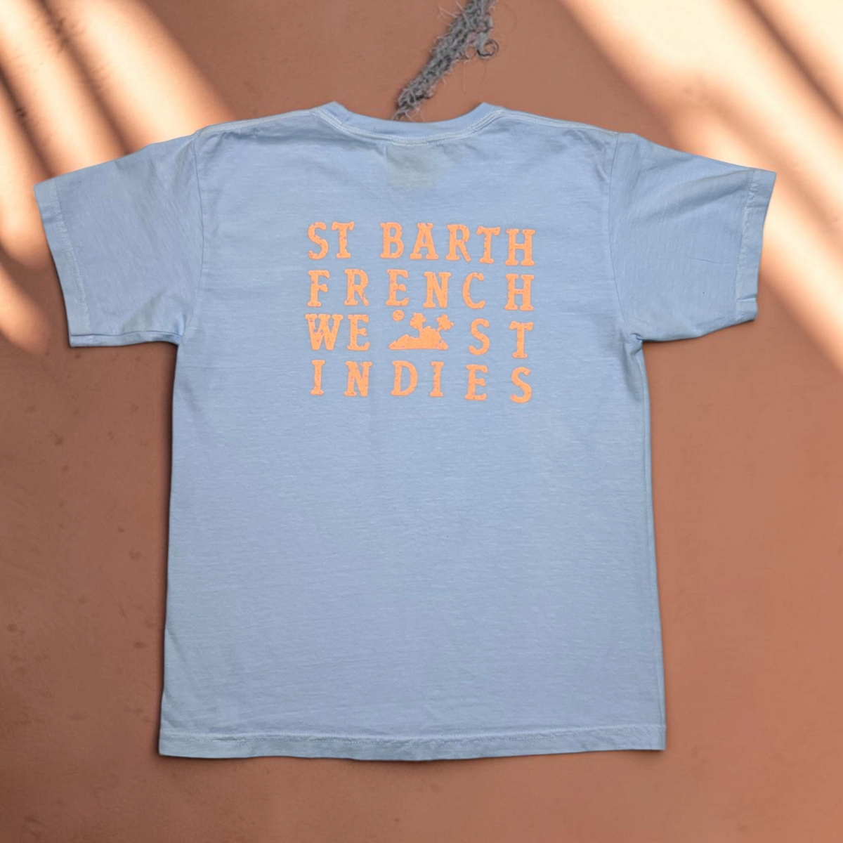 T-shirt bleu clair pour enfant St Barth French West Indies avec logo orange imprimé au dos sur cintre en bois