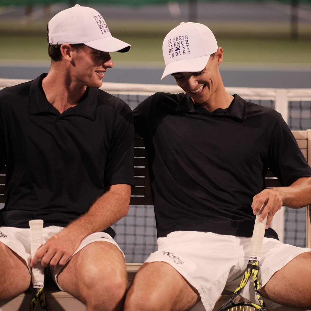 Deux hommes portant le polo noir St Barth French West Indies AUGUSTE et des casquettes blanches sur un court de tennis